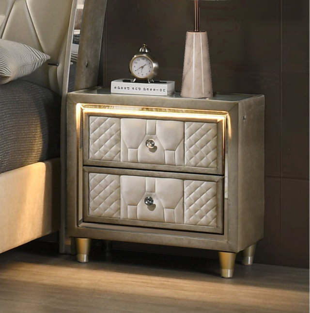 A luxury bedroom set featuring a tufted cream upholstered bed with silver trim, a matching dresser with an in-bed fire, a nightstand, and a mirror, arranged in a modern gray-toned bedroom.