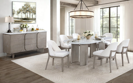 A modern dining room set featuring a round white marble-top table with a light wood pedestal base, surrounded by six white upholstered dining chairs with light wood frames. The set is styled on a large cream patterned area rug in a bright, contemporary dining room with a matching gray wood credenza and a large window in the background.